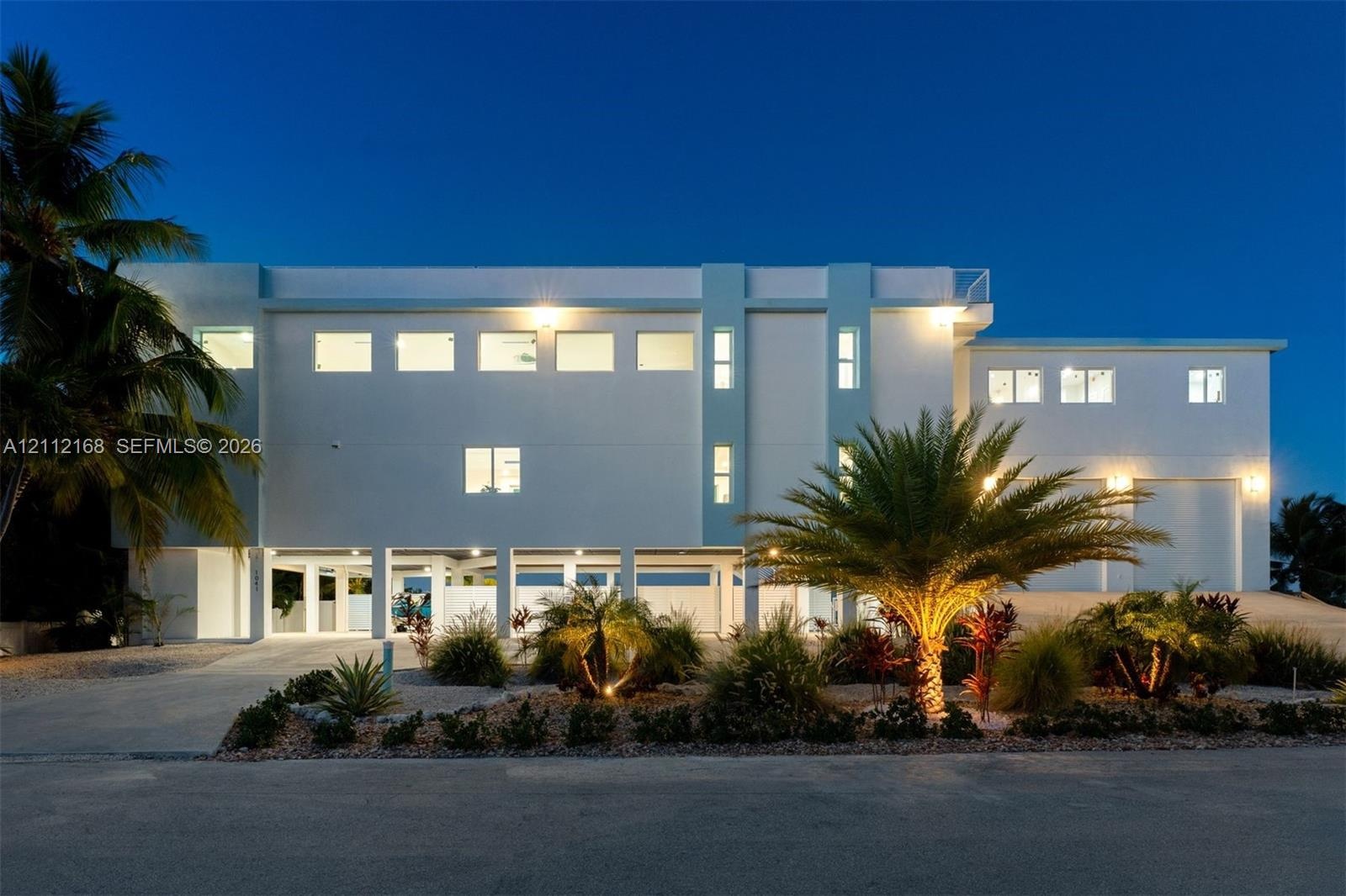 Multi-level contemporary exterior featuring a smooth stucco facade, elevated structural pilings, integrated garage bays, and a rooftop terrace