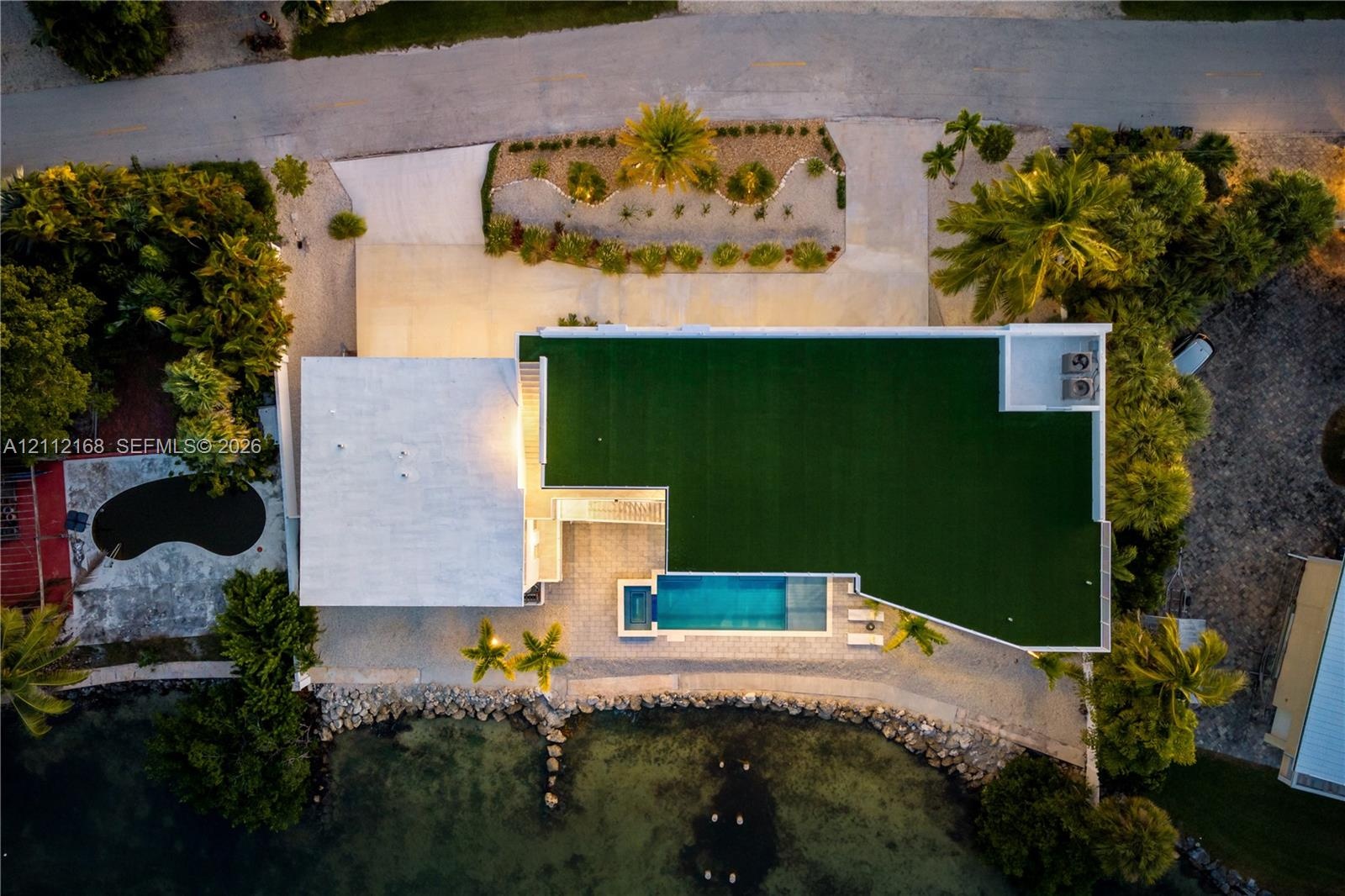 Aerial perspective featuring a waterfront property with an in-ground pool, concrete paved driveway, and established landscaping