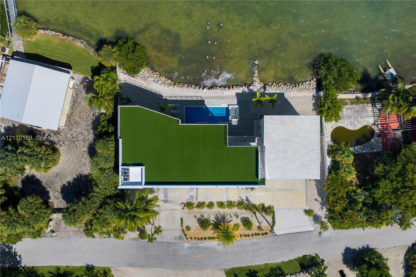 Aerial perspective of waterfront property featuring a swimming pool, synthetic turf lawn, stone-reinforced shoreline, and paved driveway