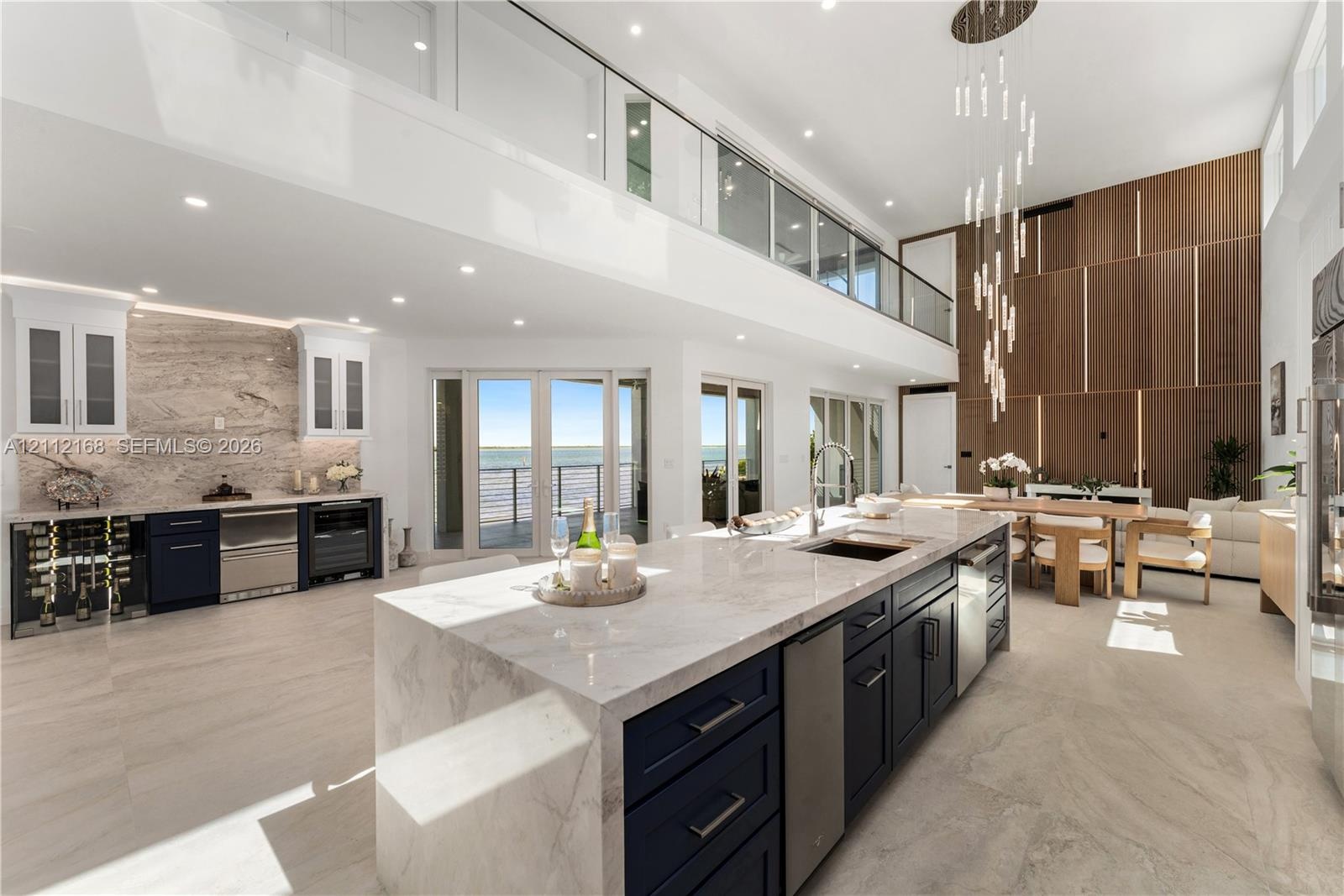 Double-height ceilings with custom wood-panel wall feature, oversized kitchen island with stone waterfall countertop, integrated floor-to-ceiling glas