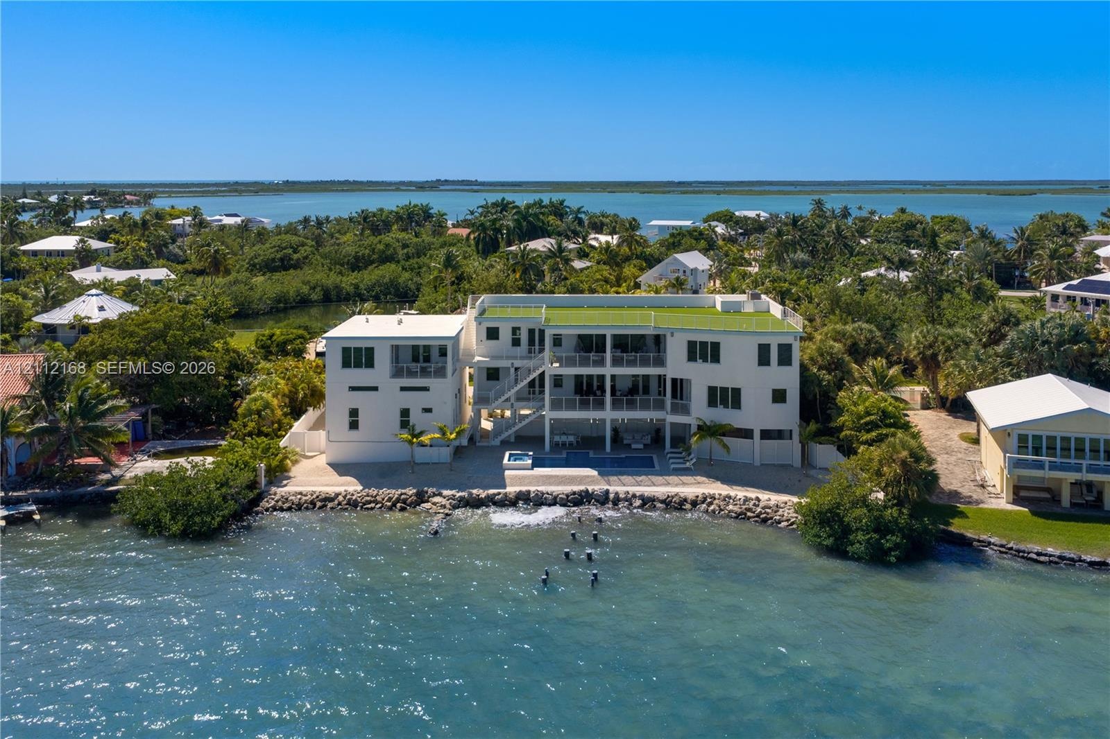 Waterfront multi-story residence featuring a ground-level swimming pool, expansive elevated balconies, stone-reinforced shoreline, and a rooftop deck