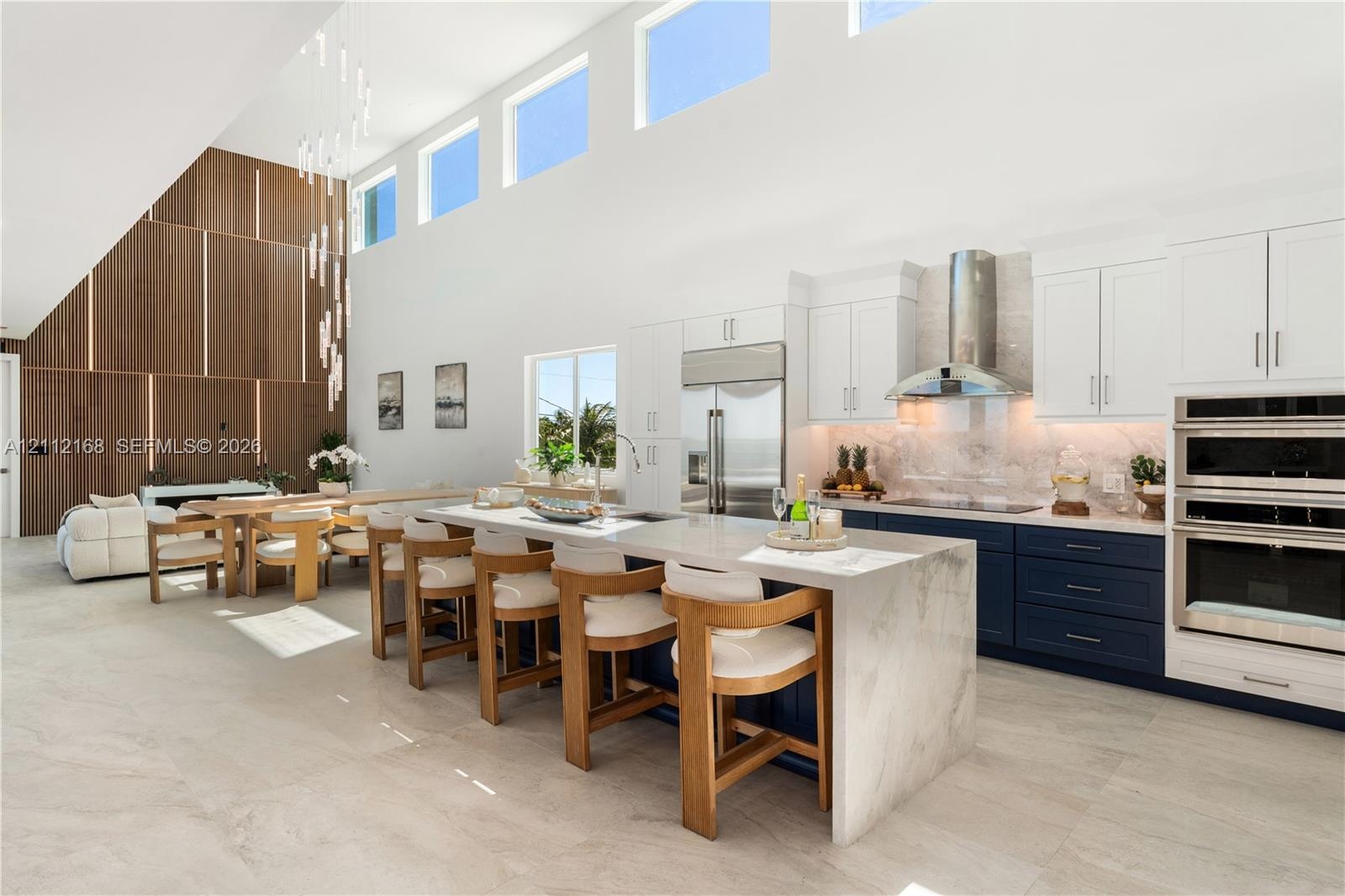 Open-concept kitchen featuring double-height ceilings, a large stone-topped waterfall island, custom two-toned cabinetry, built-in stainless steel app