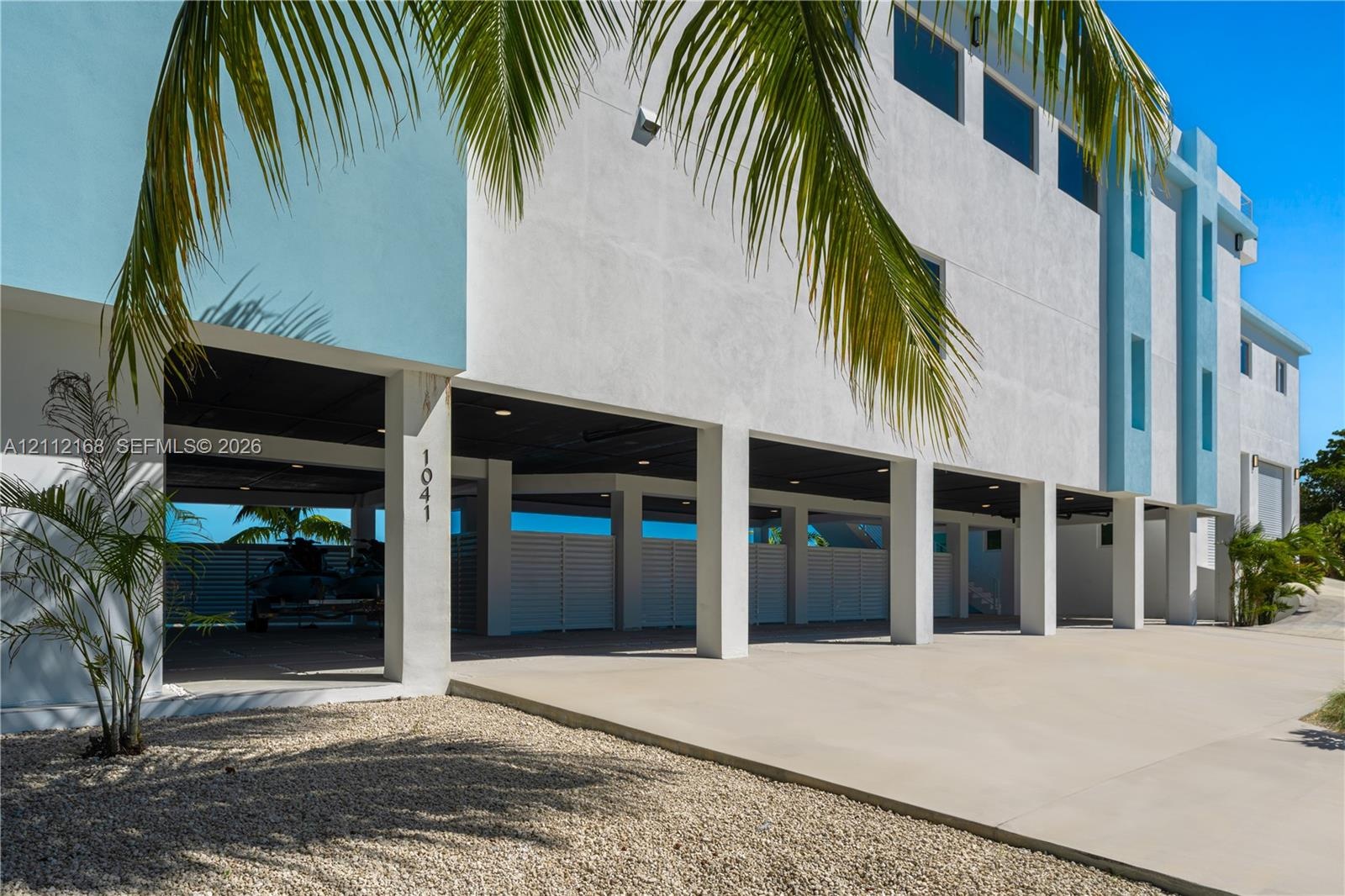 Modern multi-level exterior featuring white stucco finish, covered parking area, concrete driveway, horizontal slat privacy fencing, and recessed ligh