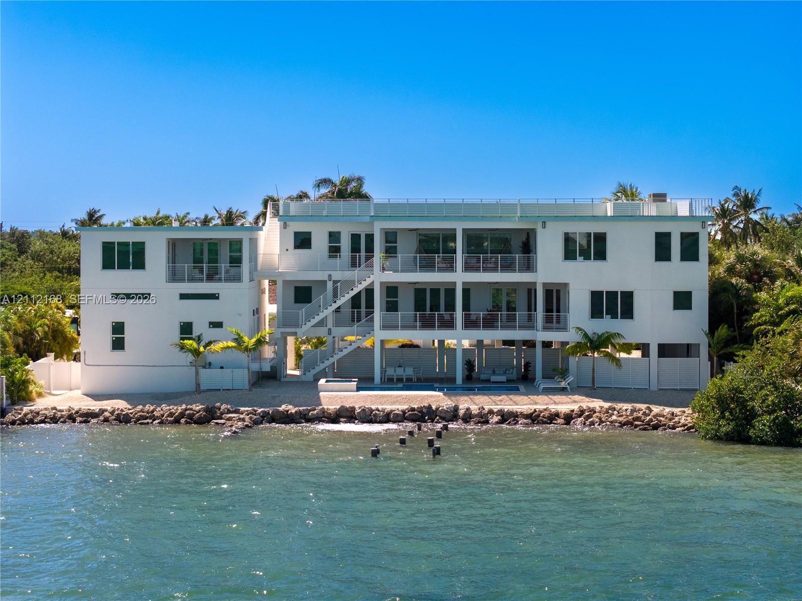 Multi-level waterfront structure featuring a white stucco exterior, tiered balconies with metal railings, an integrated ground-level swimming pool, an