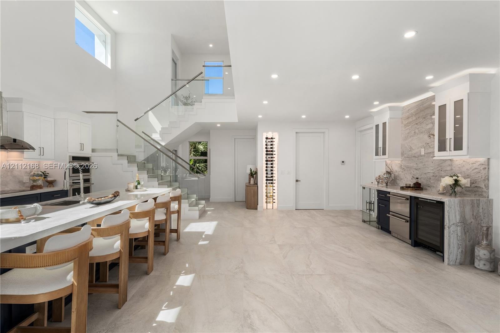 Open-concept kitchen featuring a large central island with waterfall-edge countertop, natural stone-style backsplash, integrated wine refrigeration, b