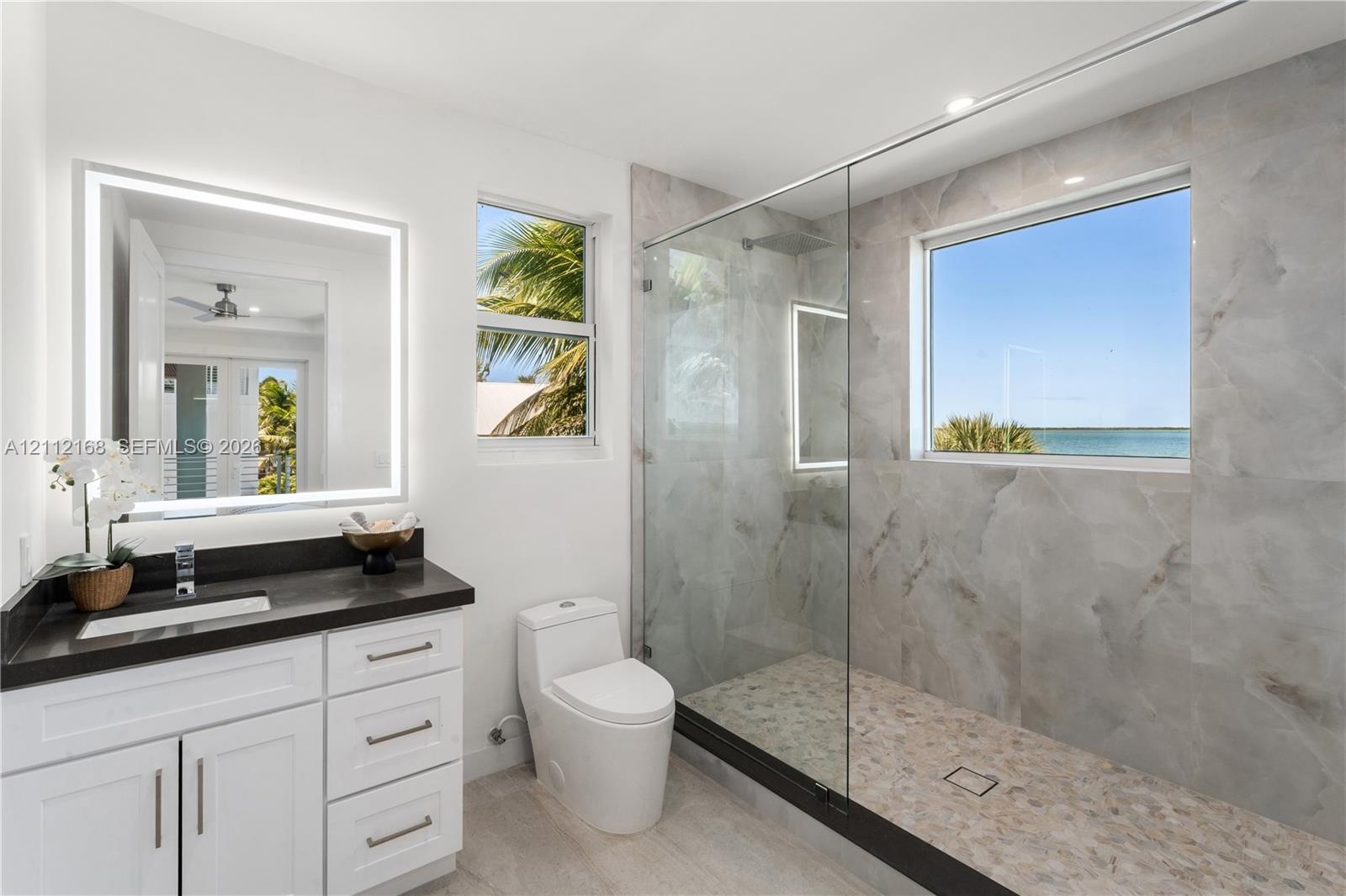 Bathroom featuring a white vanity with a dark countertop, an illuminated mirror, a glass-enclosed shower with large-format tile surround, and an integ