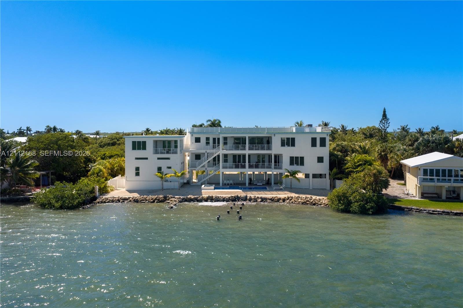 Waterfront multi-story residence featuring tiered exterior balconies, a ground-level swimming pool, and stone-reinforced shoreline