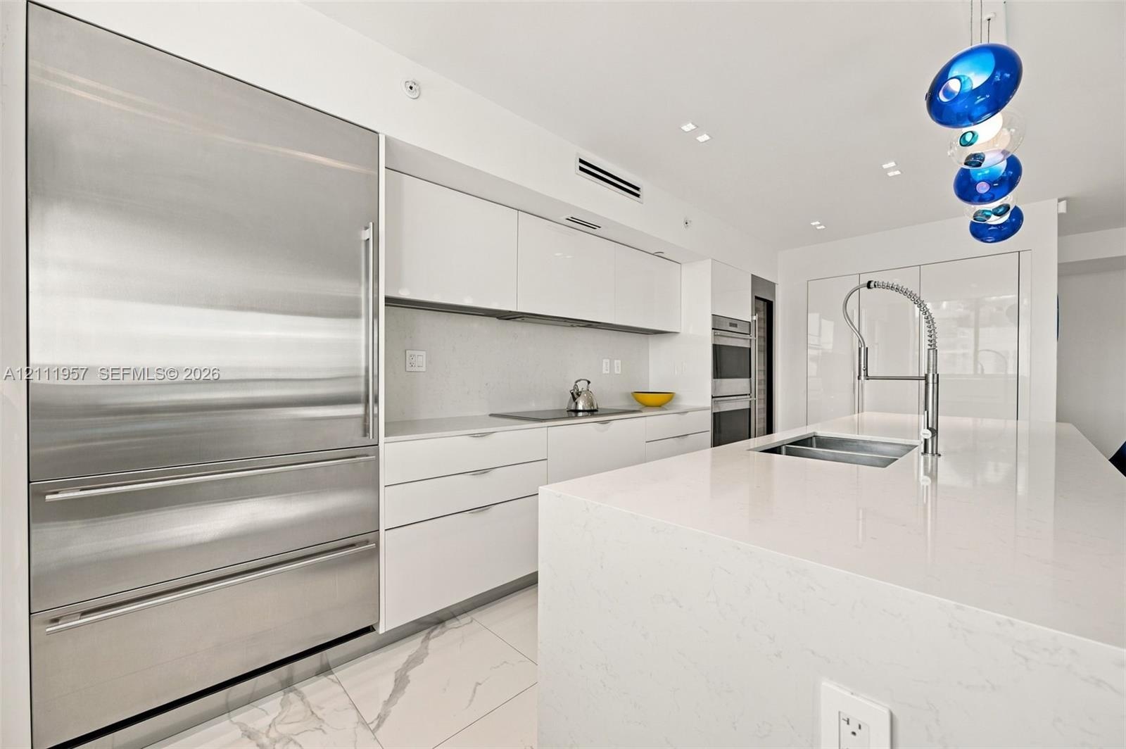 Modern kitchen featuring high-gloss cabinetry, a quartz waterfall island, stone-finish flooring, integrated stainless steel refrigeration, and a comme