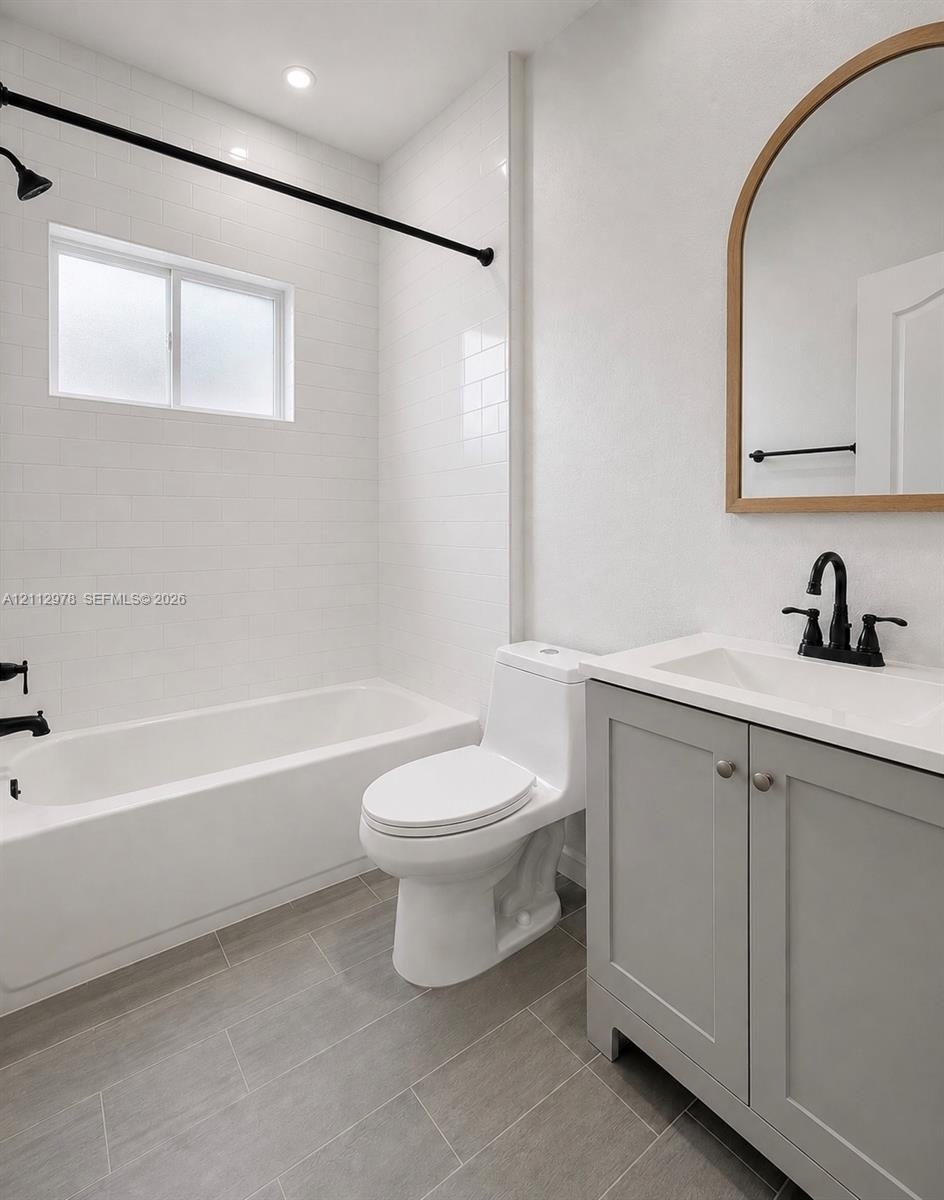 Bathroom featuring a grey vanity with a white countertop, matte black fixtures, an arched wood-frame mirror, tile shower surround, and grey flooring