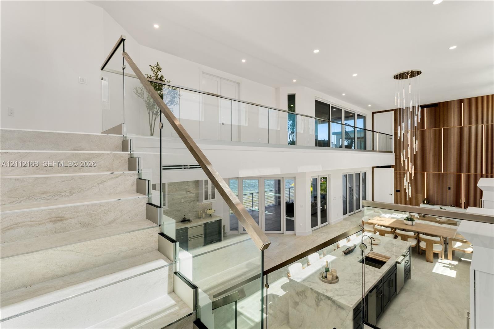 Open-concept interior featuring stone-finish flooring, a glass and metal staircase railing, a multi-level wood-panel wall feature, a stone-topped kitc