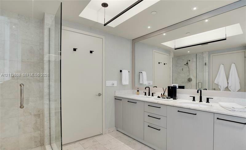 Dual-sink vanity with light-gray cabinetry, stone countertops, matte black fixtures, full-width mirror, and walk-in tiled shower with a glass enclosur