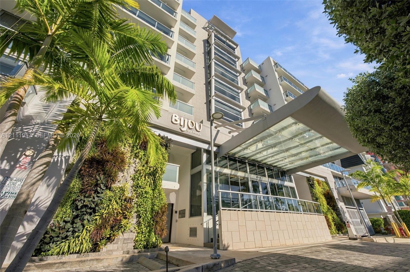 Multi-story residential building exterior featuring a glass-canopy entryway, tiered balconies with glass railings, vertical gardens, and stone-tiled f