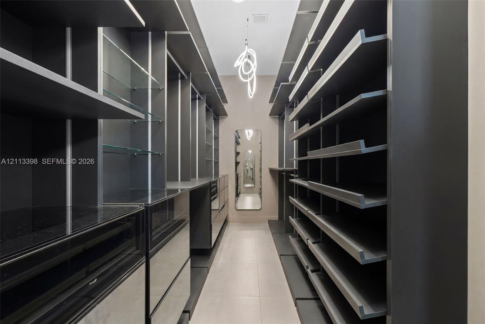 Custom walk-in storage area featuring dark-toned built-in cabinetry, integrated glass shelving, sleek flat-front drawers, a full-length wall mirror, a