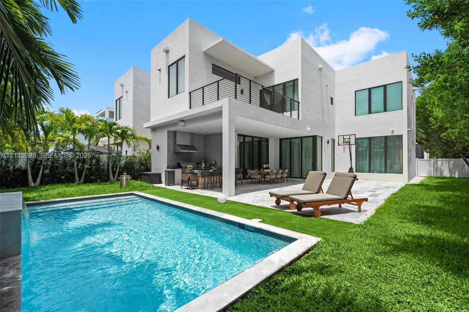 Contemporary residence featuring a stucco exterior, second-story balcony with metal railing, covered outdoor kitchen, stone-paved patio, and a swimmin