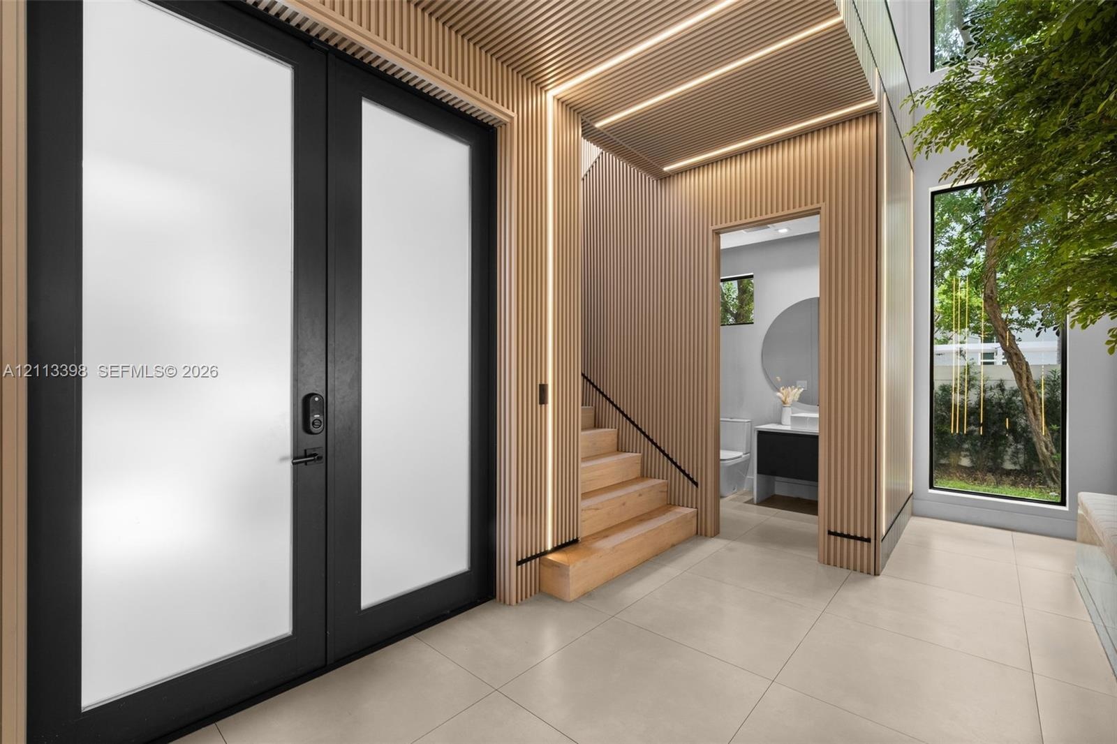 Double-entry door with frosted glass panels, wood-slat wall paneling with integrated LED lighting, large-format tile flooring, and a floating staircas