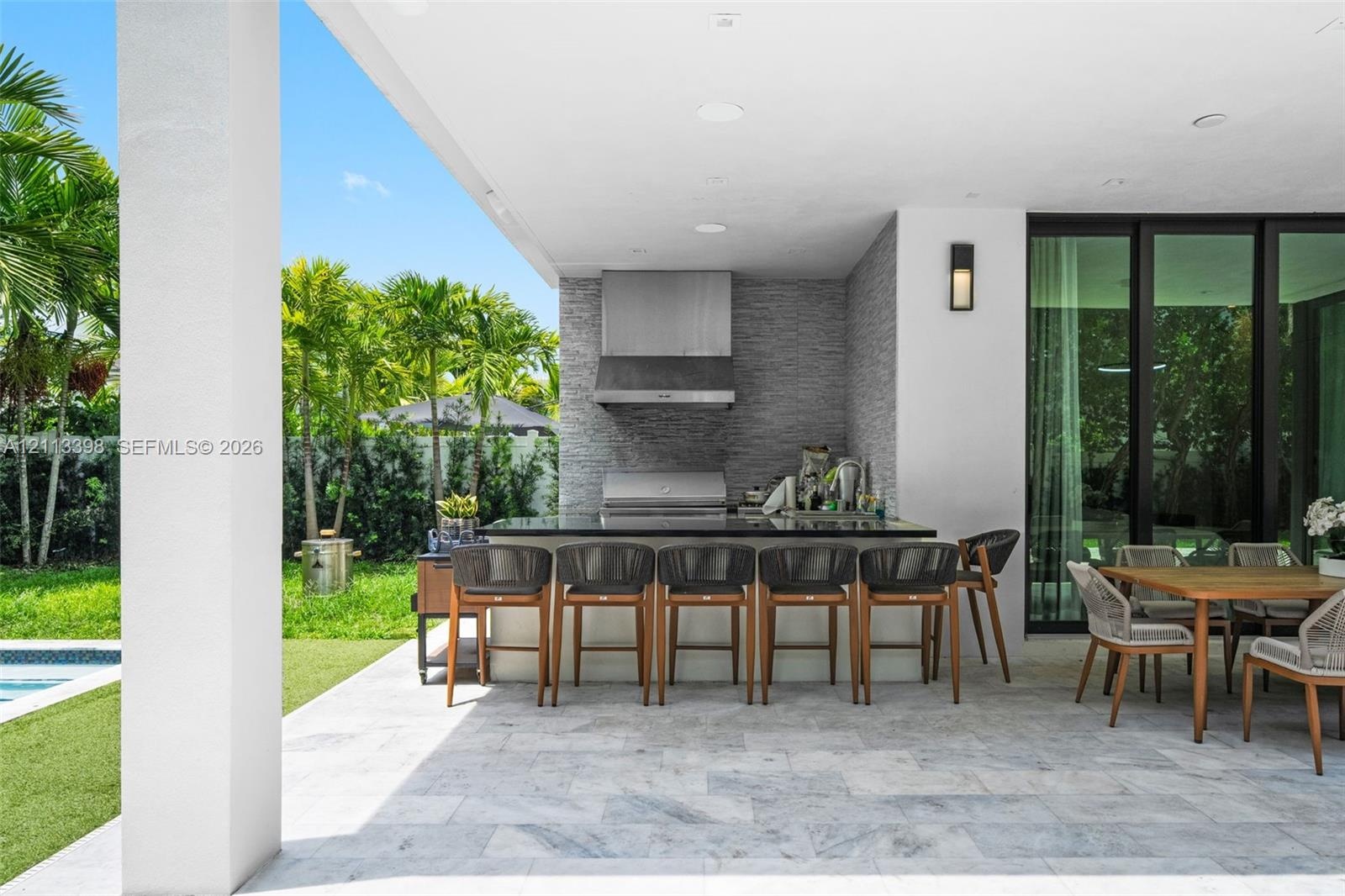 Covered outdoor patio featuring light-toned stone tile flooring, a built-in outdoor kitchen with a stainless steel range hood, natural stone backsplas