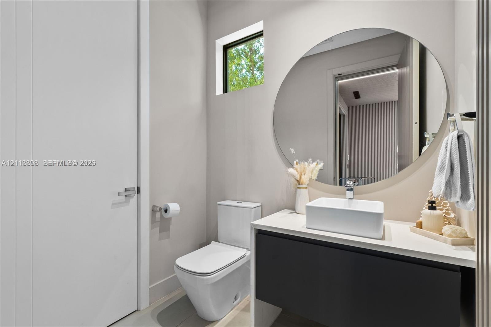 Floating dark-finish vanity with a white rectangular vessel sink, solid-surface countertop, circular frameless mirror, and dual-flush toilet