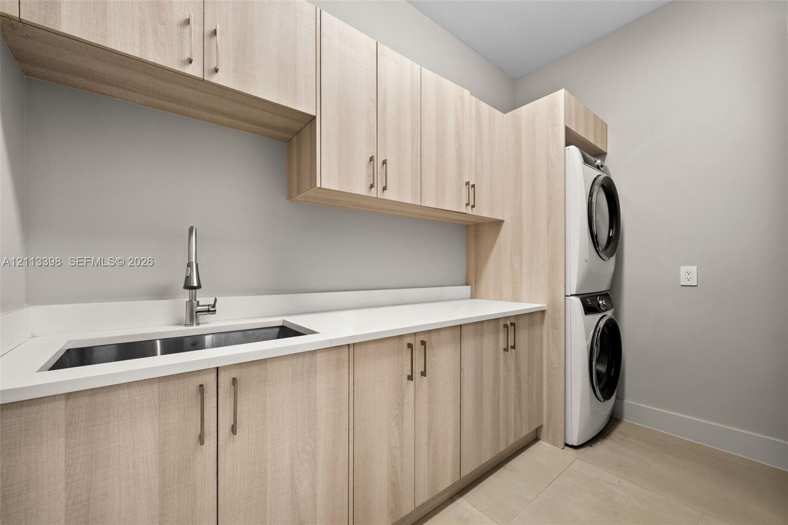 Laundry room featuring light wood-finish cabinetry, white solid-surface countertops, an undermount stainless steel sink with a high-arch pull-down fau