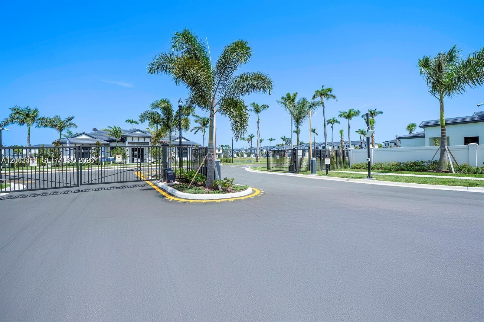 Gated community entrance featuring metal perimeter fencing, paved roadway, manicured landscaping, and mature palm trees