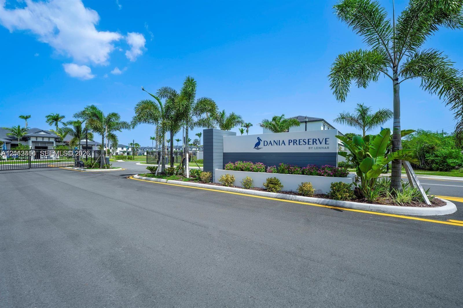 Gated community entrance featuring a masonry monument sign, asphalt paved roadway, and tropical landscaping