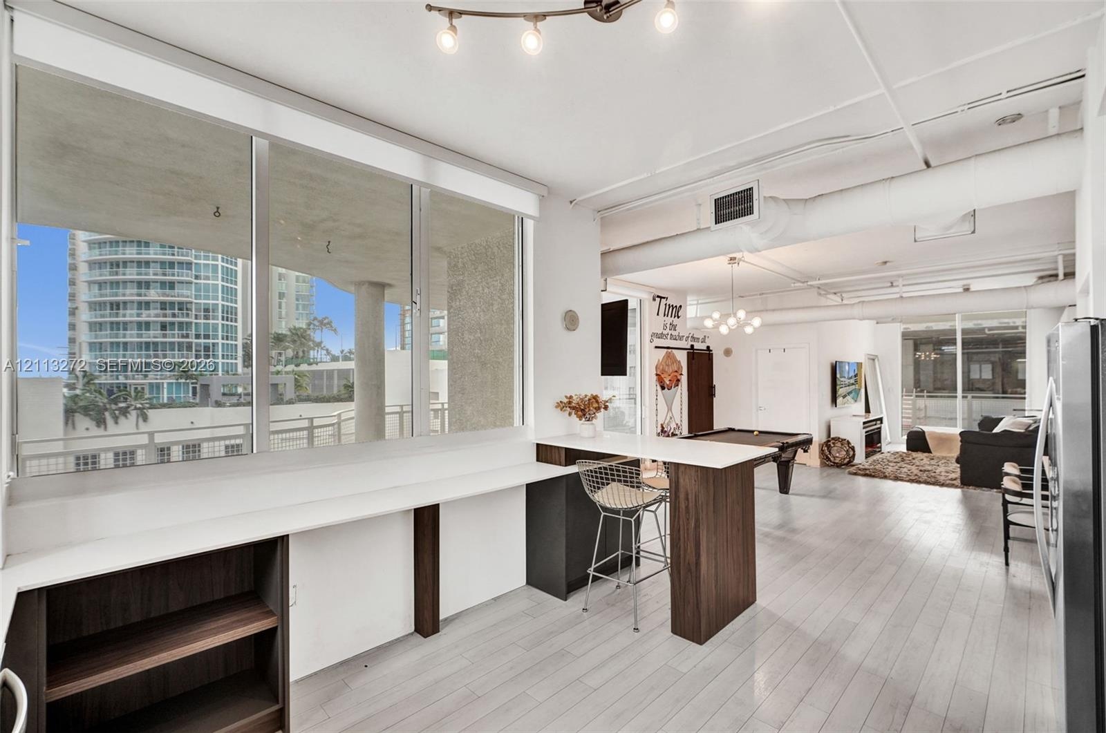 Expansive floor-to-ceiling windows, white-toned wood-finish flooring, industrial-style exposed ductwork, and built-in breakfast bar featuring a sleek 