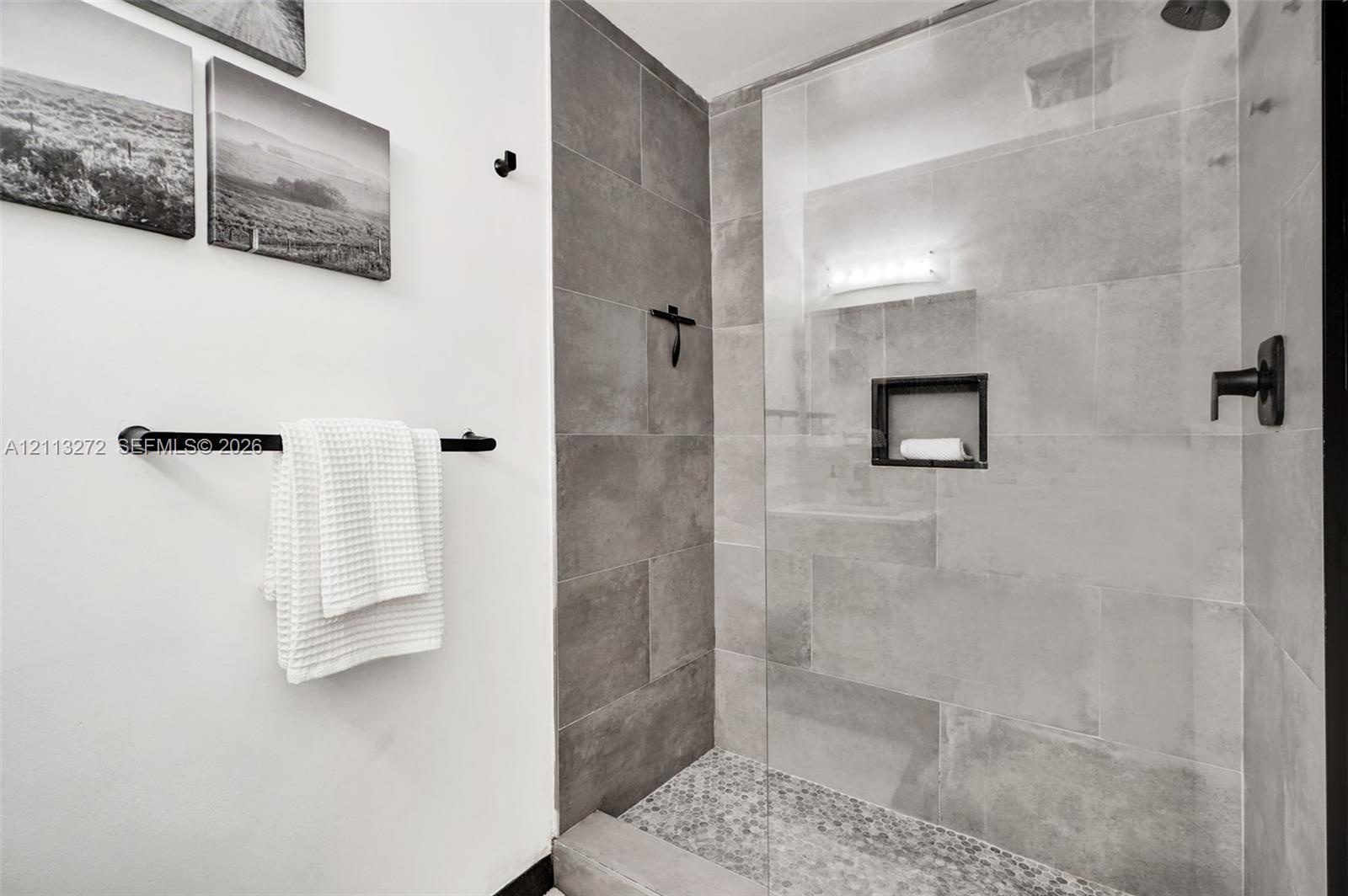 Walk-in shower featuring large-format grey tile surround, pebble-texture floor, frameless glass enclosure, built-in recessed wall niche, and matte bla