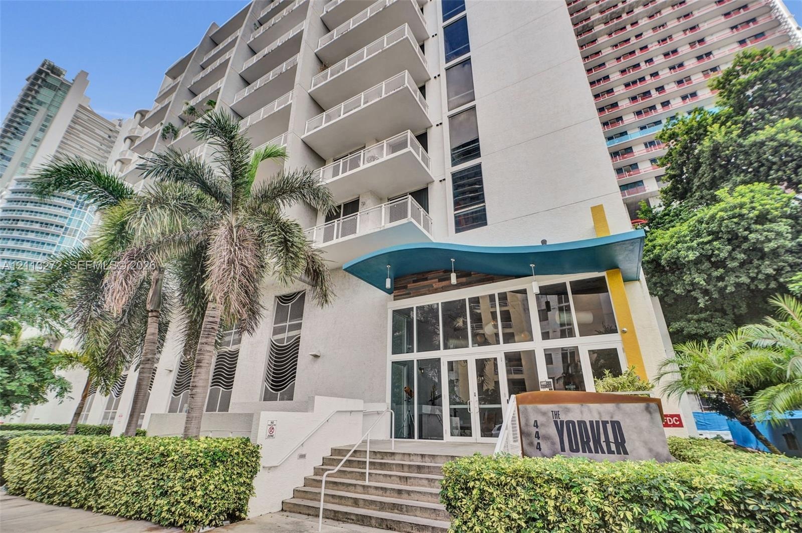 Multi-story high-rise exterior featuring concrete balconies, floor-to-ceiling glass-paneled ground-level entryway, decorative blue architectural awnin