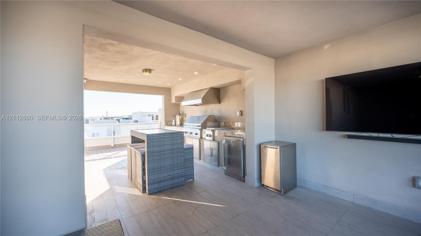 Covered outdoor kitchen featuring a stainless steel grill, range hood, under-counter refrigeration, and stone-finish countertops, complemented by tile