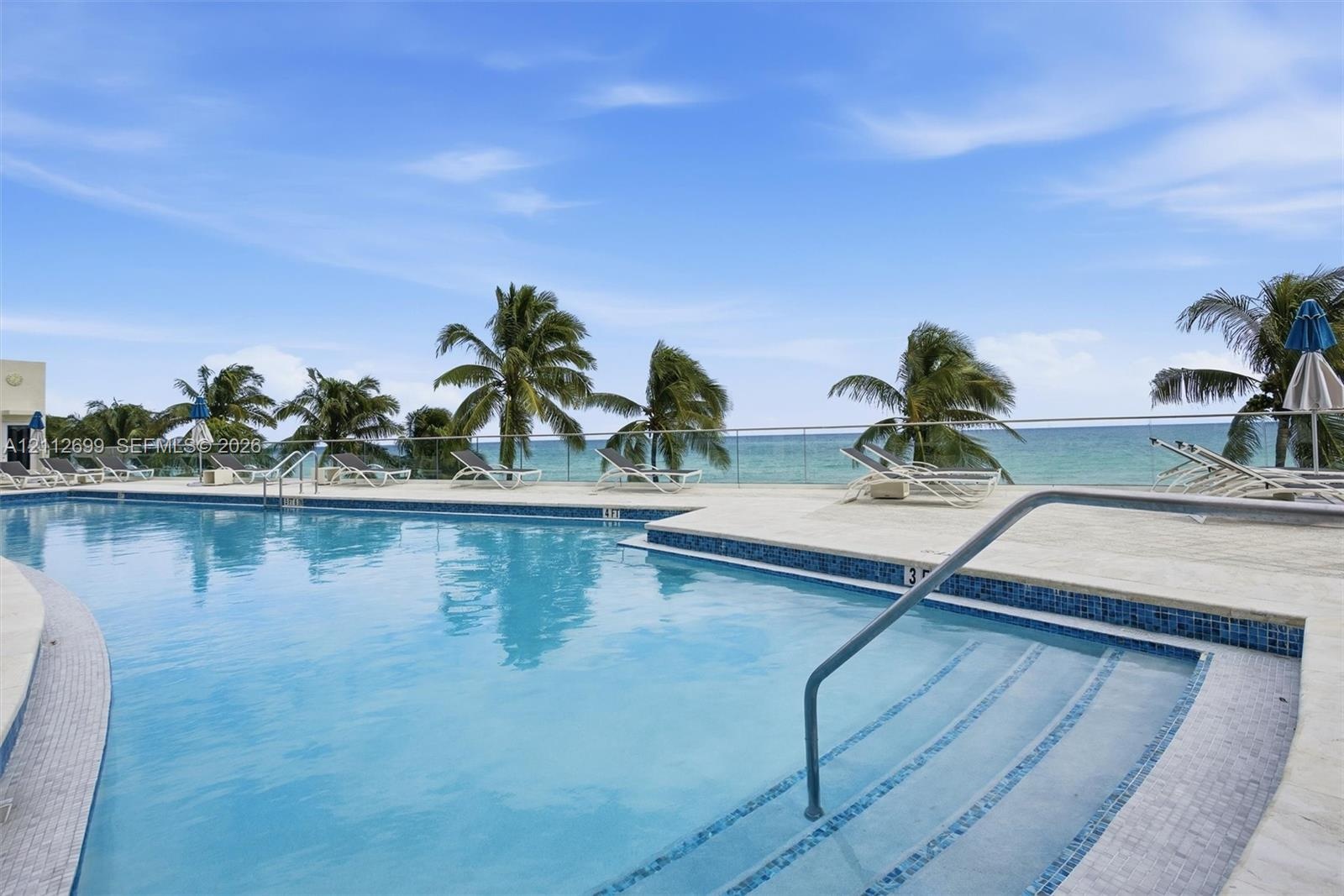 Swimming pool with mosaic tile border, light-toned stone decking, metal handrail, and transparent glass railing overlooking the ocean