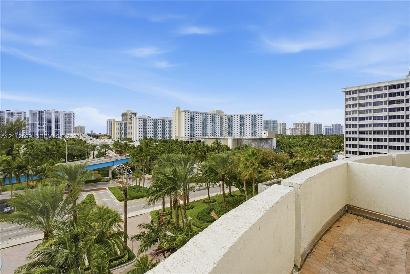 Balcony with textured masonry walls and tiled flooring overlooking an urban landscape featuring mature palm trees and multi-story structures