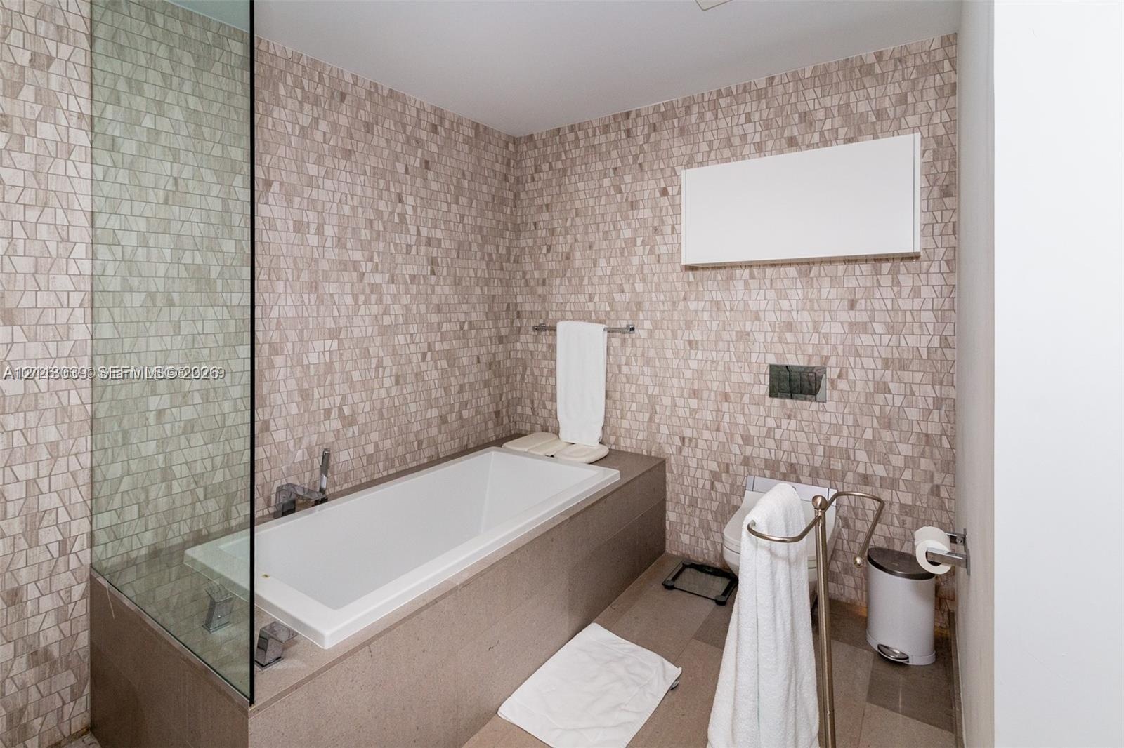 Full-height mosaic tile wall surfacing, deep rectangular soaking tub with deck-mounted chrome faucet, glass panel enclosure, and wall-mounted water cl