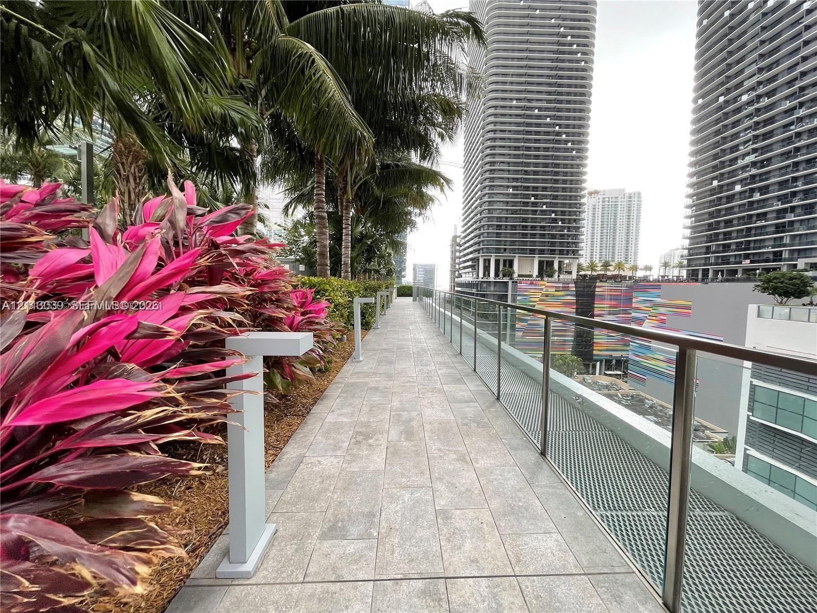 Terrace featuring large-format stone-look tile flooring, glass panel railing, and integrated metal path lighting, bordered by mature palm trees and tr