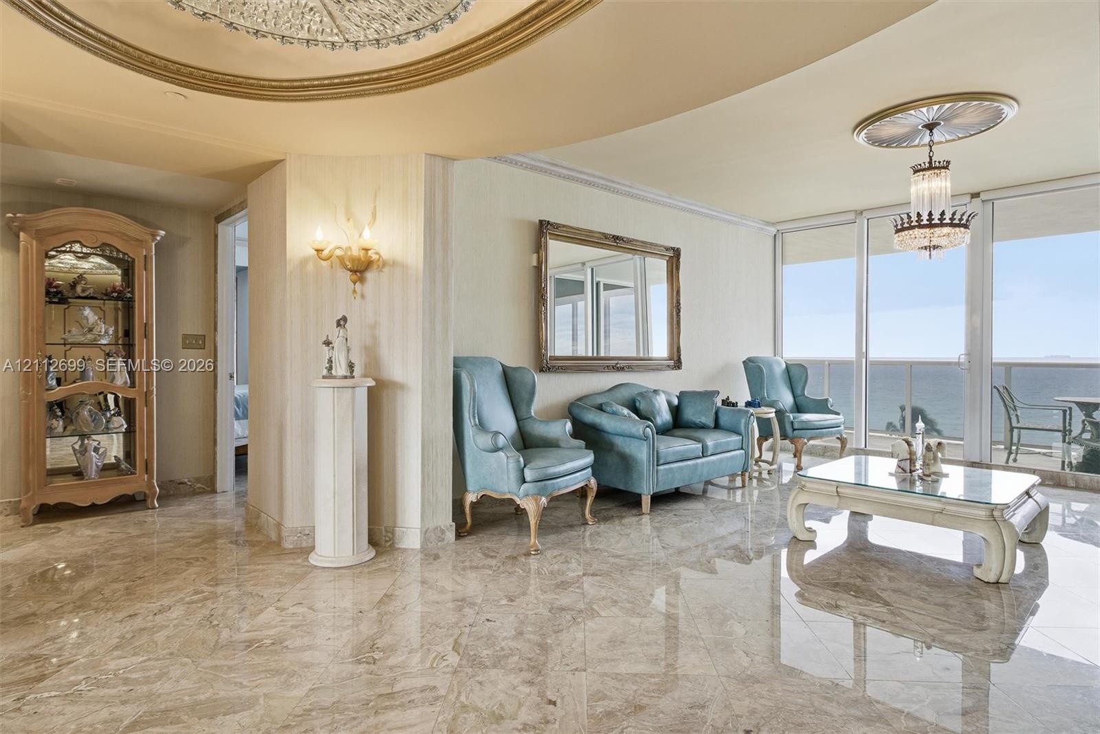 Polished stone flooring, recessed tray ceiling with gold-tone molding, crystal chandelier, and floor-to-ceiling glass sliding doors with ocean backdro