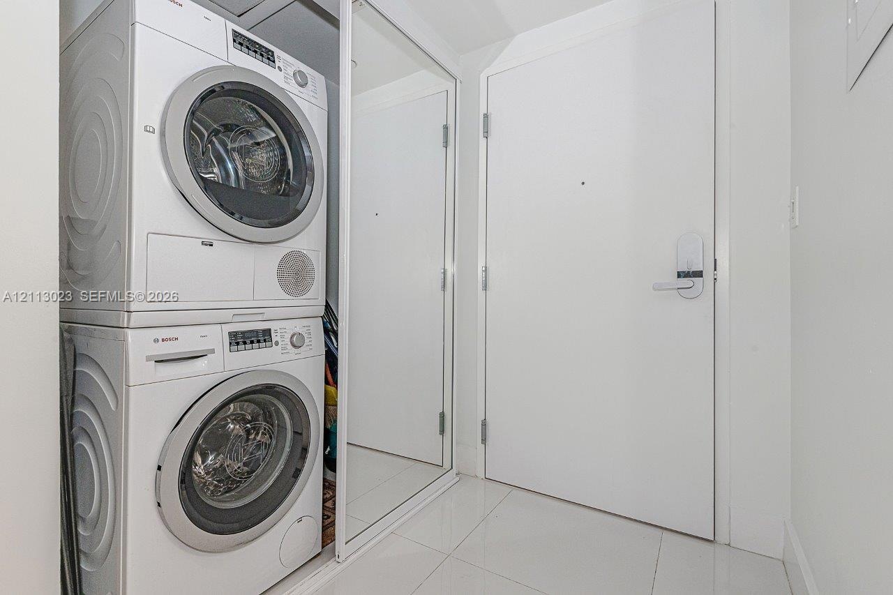 Stacked Bosch laundry center enclosed by sliding mirrored doors, set against large-format tile flooring and a solid-core door featuring electronic key