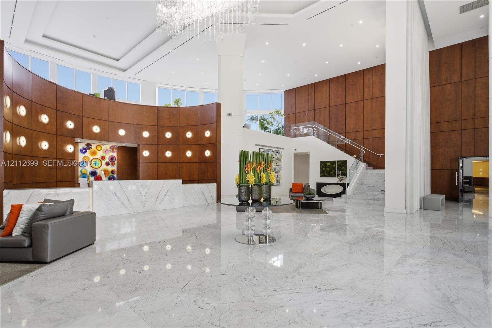 Grand entrance featuring high ceilings, polished stone flooring, curved wood-paneled walls, a statement crystal chandelier, and a built-in stone recep