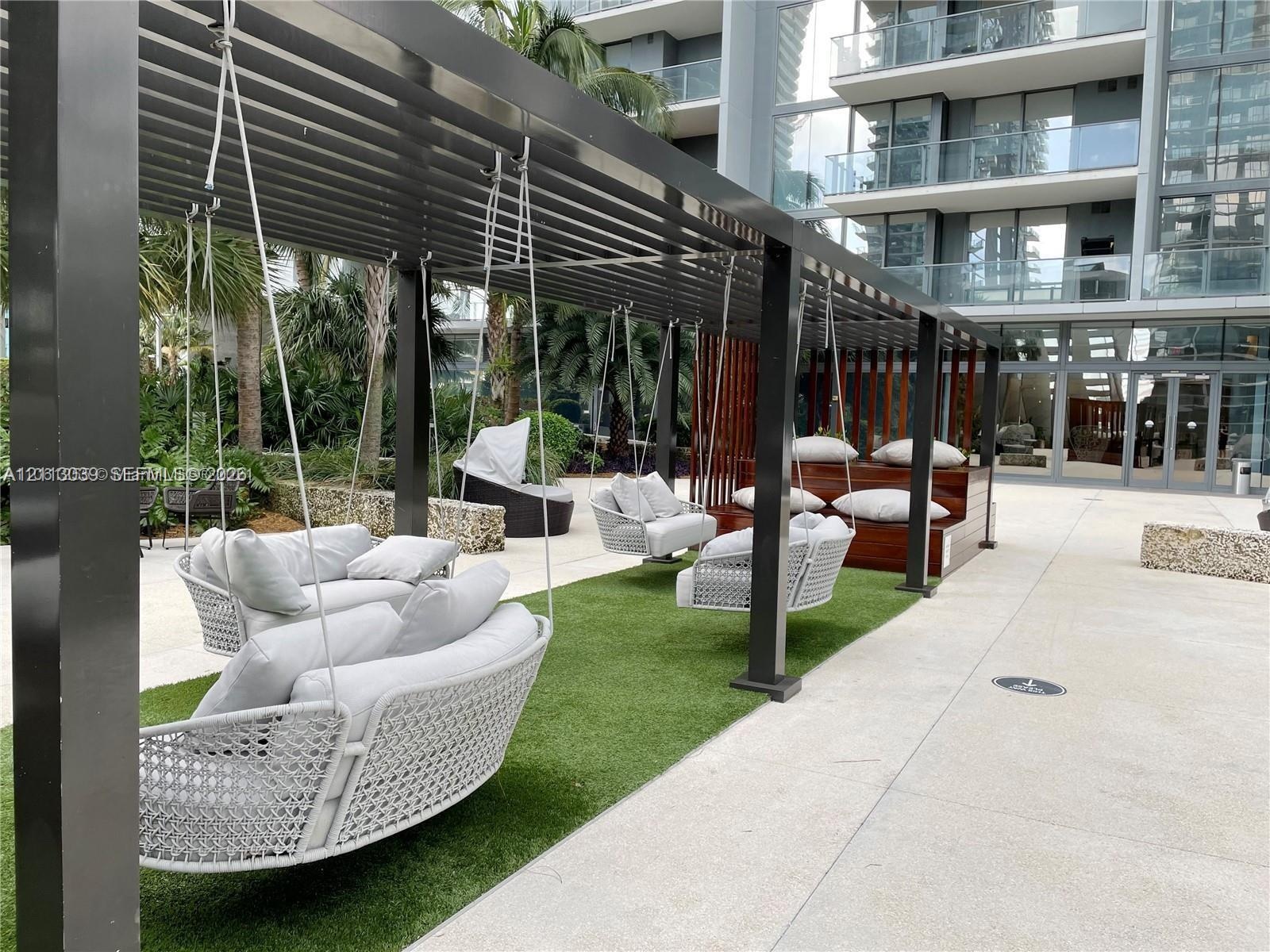 Outdoor common area featuring a black metal pergola, integrated artificial turf flooring, built-in tiered wood-tone seating, and suspended seating str