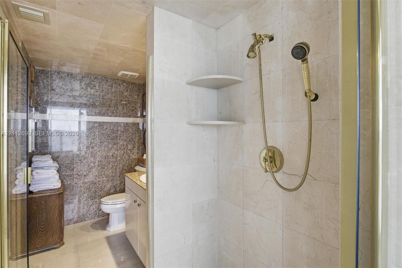 Walk-in shower enclosure featuring floor-to-ceiling neutral tile, built-in shelving, and gold-finish hardware including a handheld showerhead