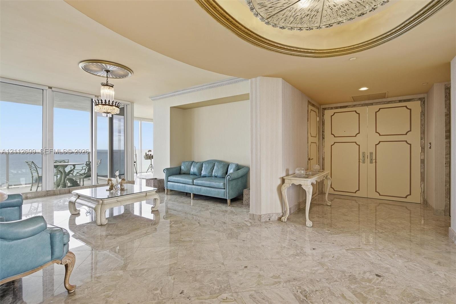 Polished stone flooring, recessed ceiling with gold-leaf detailing, dual-panel entry door with gold trim, crystal chandelier, and wall-to-wall glass s