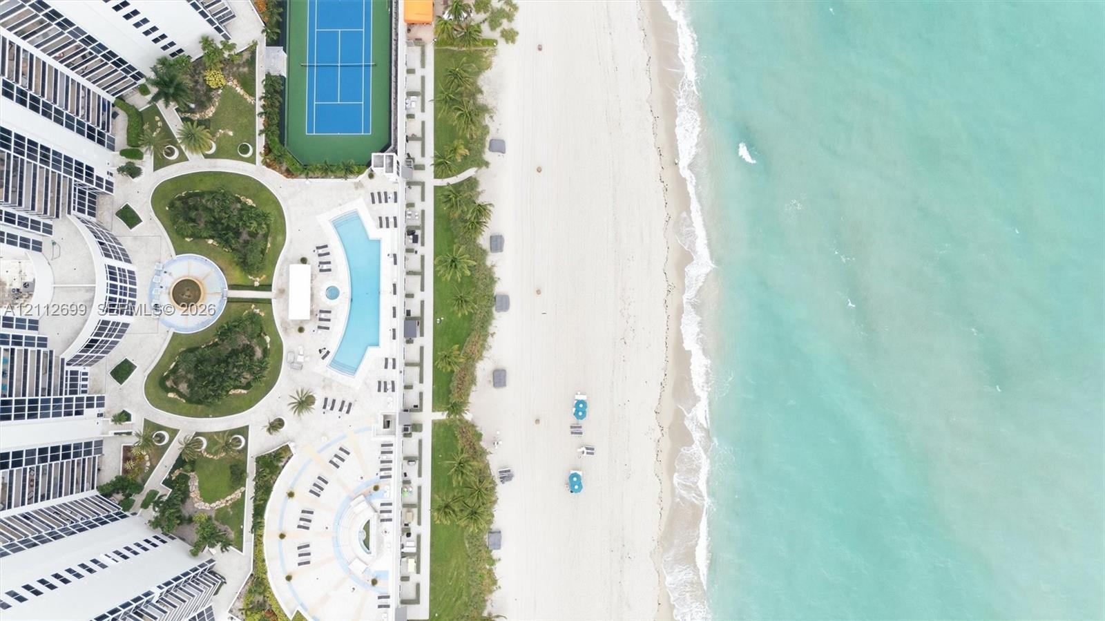 Aerial perspective of a coastal property featuring direct beach access, a swimming pool, a tennis court, and manicured landscaping