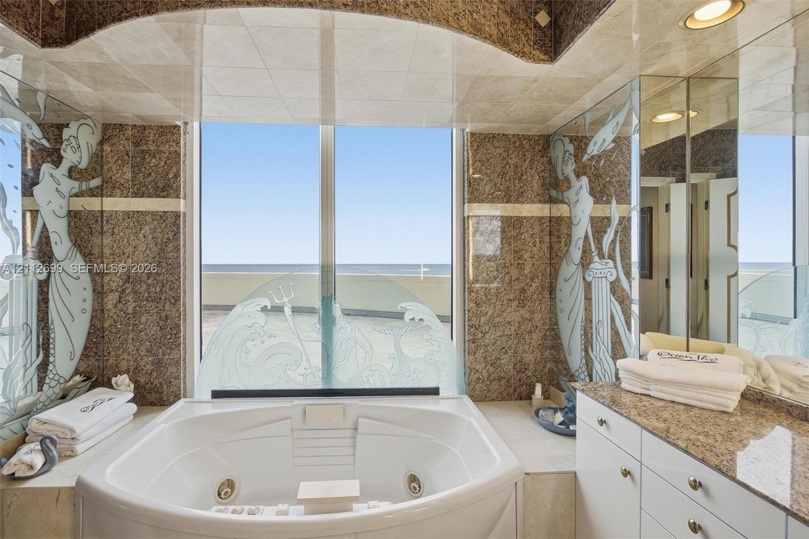 Custom soaking tub with integrated jets, large window with ocean horizon backdrop, stone wall cladding, decorative etched glass panels, and built-in v