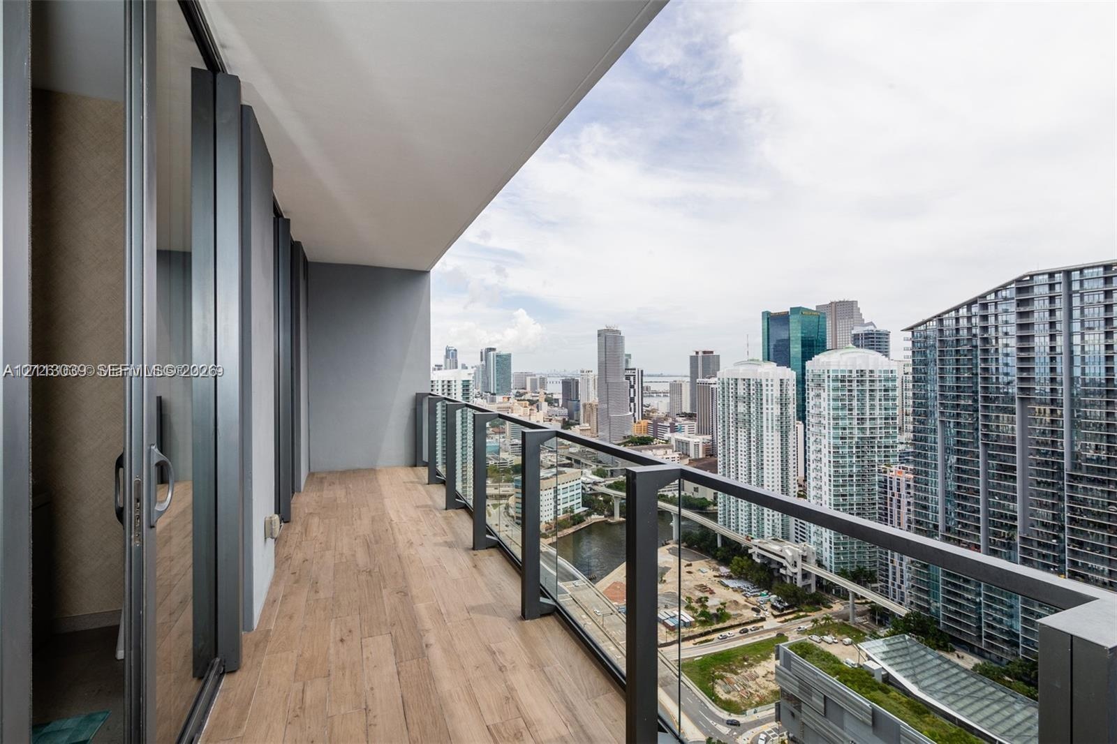 Covered balcony featuring wood-finish flooring, glass panel railing, and metal framing with a backdrop of urban architecture