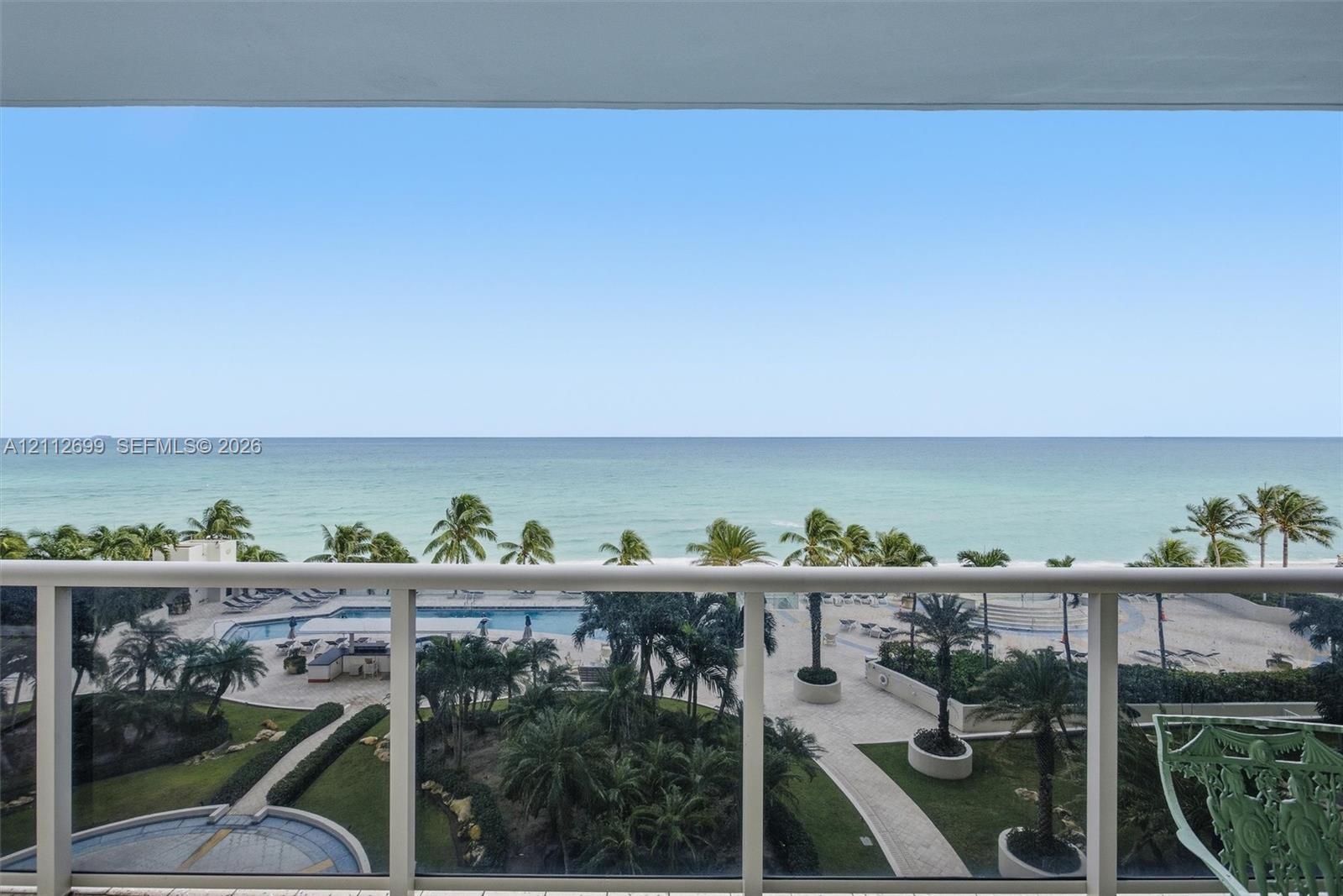 Covered balcony featuring white metal railings, overlooking an oceanfront landscape with a swimming pool, paved walkways, and palm trees