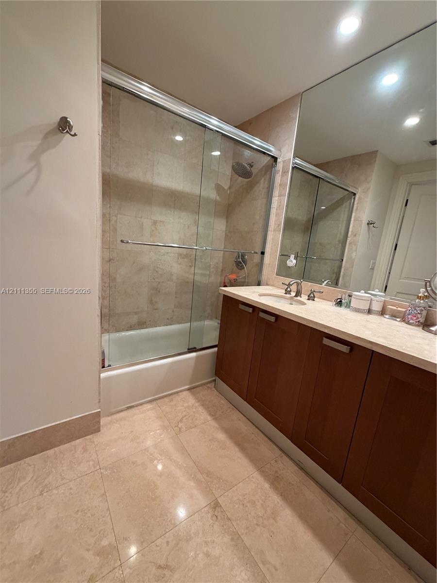 Bathroom featuring a vanity with wood-finish cabinetry, a light-toned stone-style countertop, a wall-spanning mirror, and a tub-shower combination enc