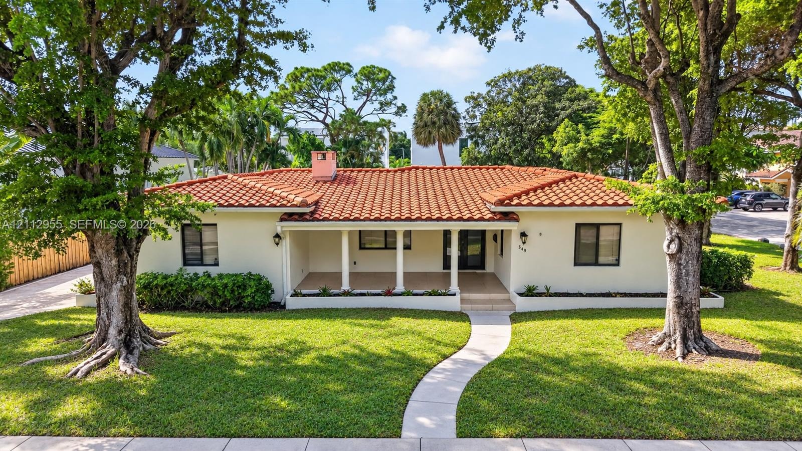 Single-story residence featuring a tile roof, covered front porch, stucco exterior, mature trees, and a manicured lawn