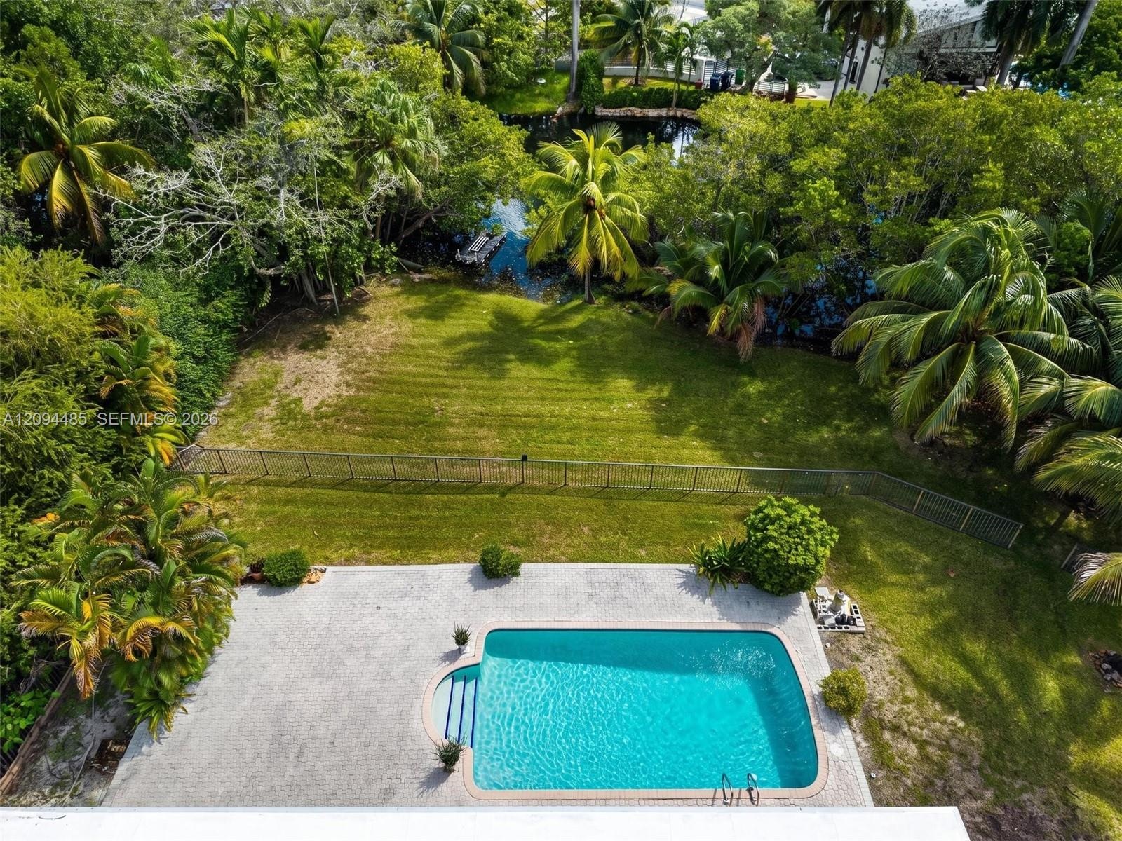Outdoor living space featuring an in-ground swimming pool, concrete paver patio, metal perimeter fencing, manicured lawn, and mature tropical landscap