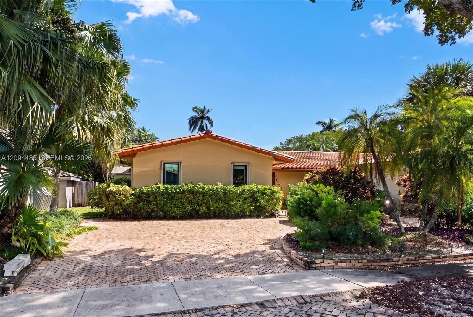 Single-story residence featuring a terracotta-tiled roof, brick-paved driveway, and lush tropical landscaping