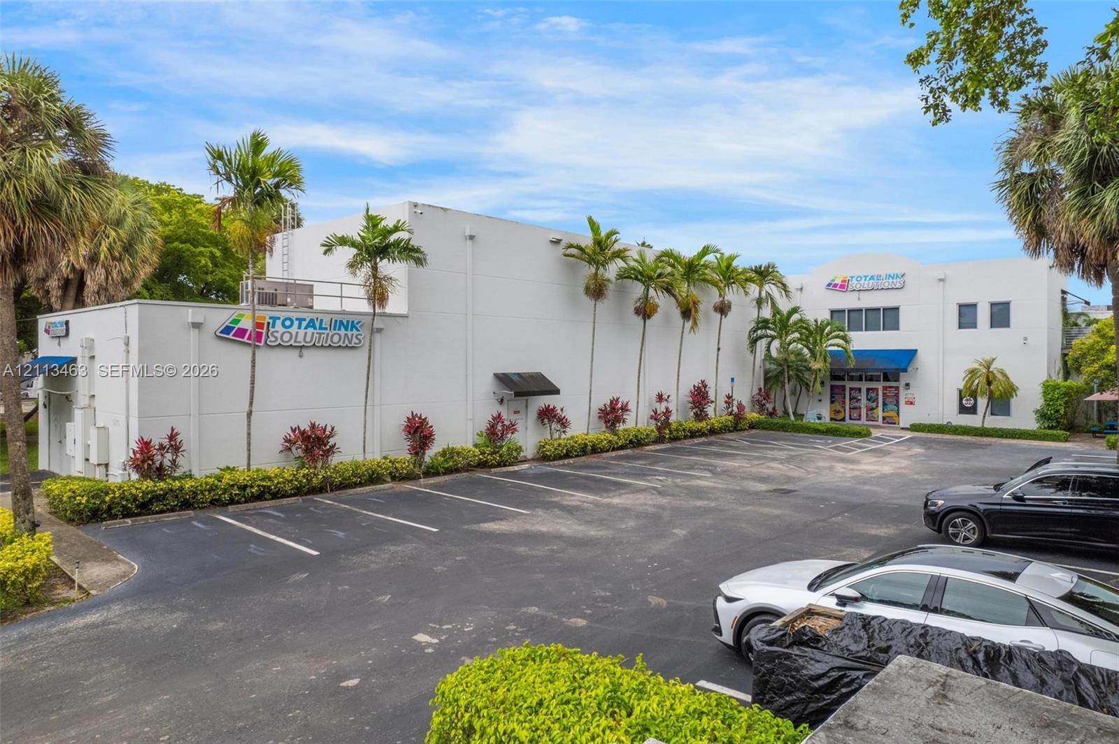 Commercial exterior featuring a concrete facade, expansive asphalt parking area, blue metal awnings, and landscaped perimeter with mature palm trees