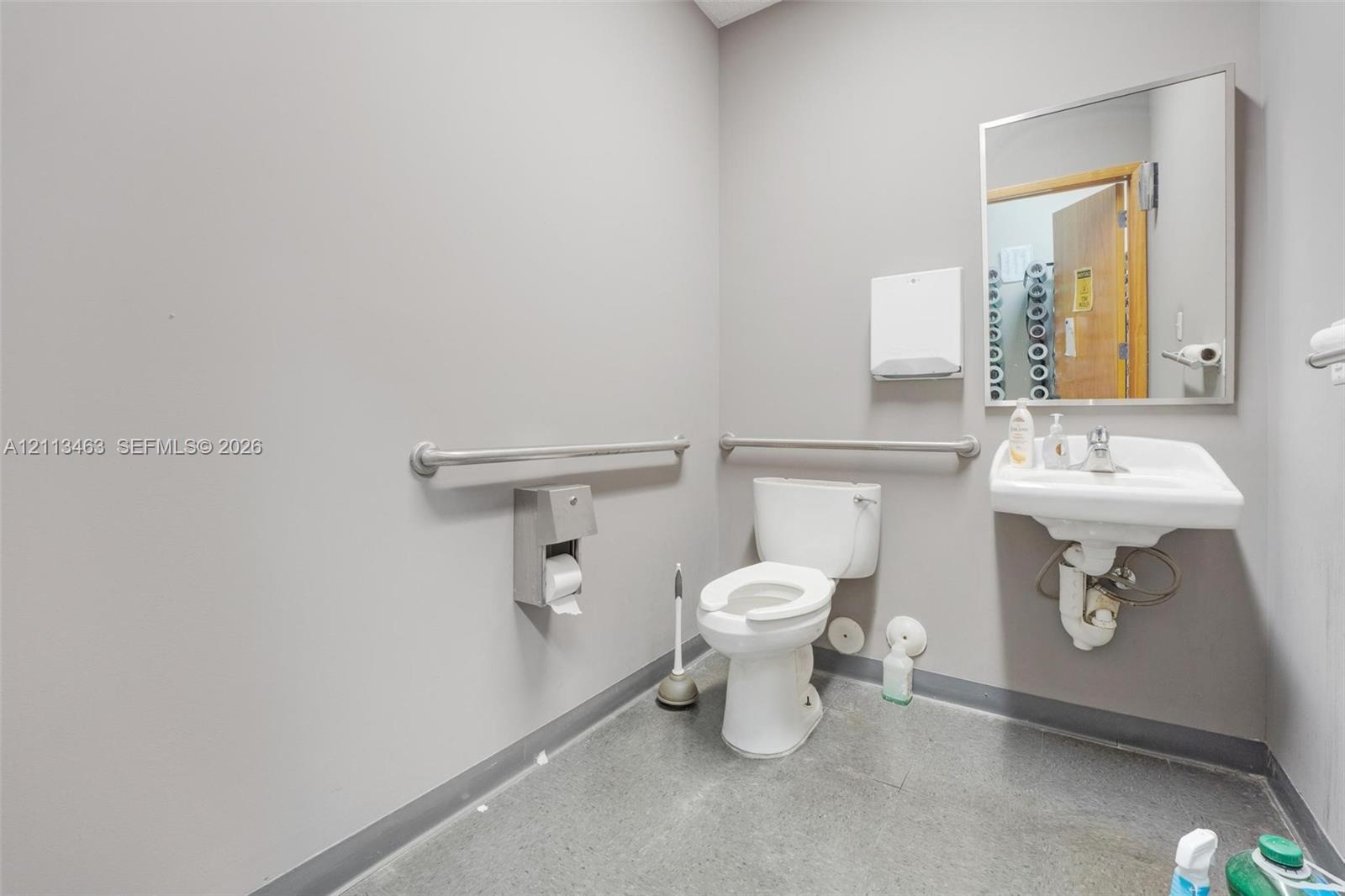 Restroom featuring a wall-mounted vitreous china sink, stainless steel grab bars, a rectangular mirror with a metal frame, and a wall-mounted paper di