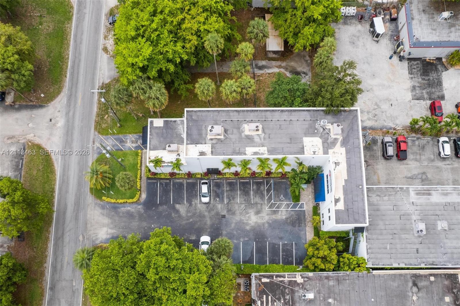 Aerial view of a commercial property featuring a dedicated asphalt parking lot, mature landscaping with established palms, and a flat roofline