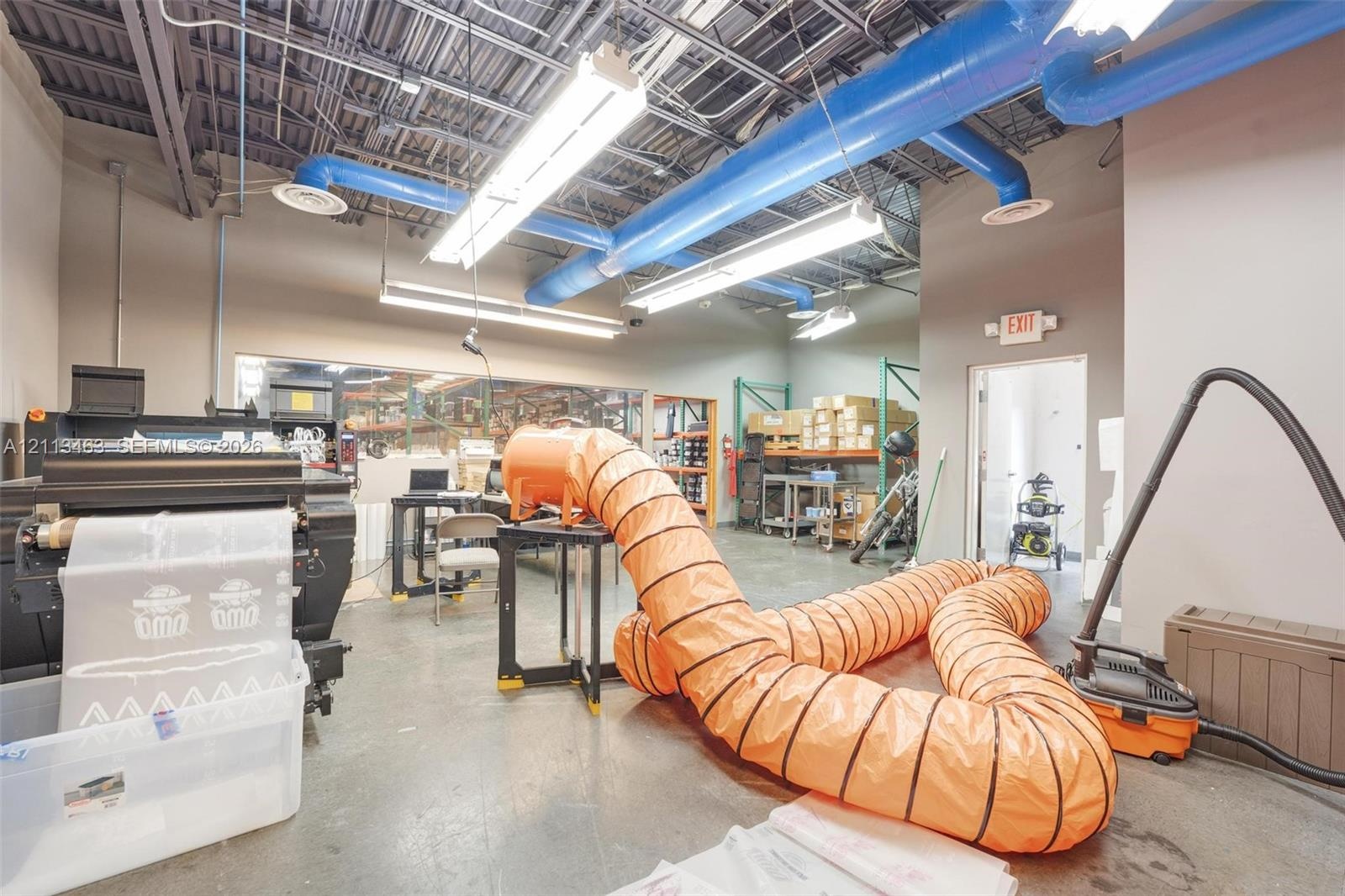 Industrial-style workspace featuring high-clearance exposed ceilings, concrete flooring, linear fluorescent lighting, and extensive blue ductwork